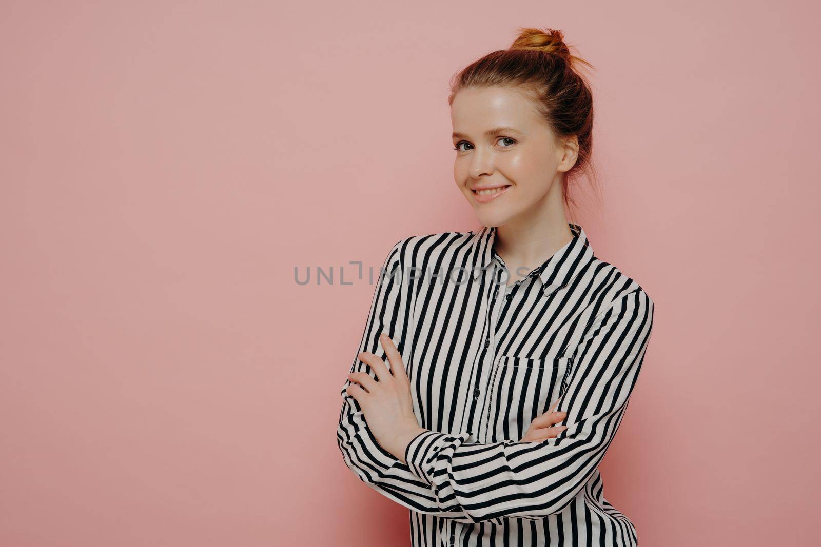 Adorable happy ginger teenage girl in striped shirt with hair in bun looking at camera with bright smile and expressing positive emotions, standing sideways with crossed arms against pink background