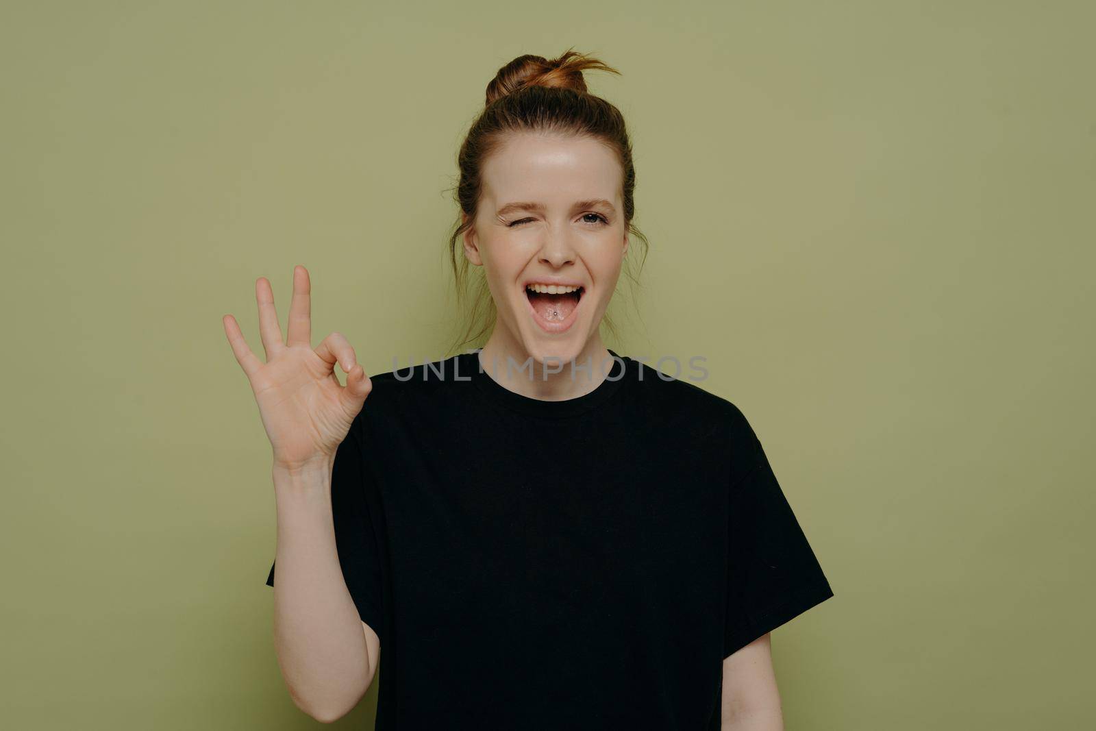 Overjoyed excited young woman in black tee shirt winking with open mouth, making ok sign with hand, assuring everything is going good and well, posing against green wall