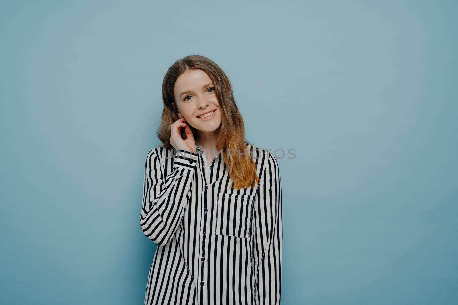 Tender cute european teen girl wearing white shirt with black stripes, expressing her shyness with tilted head and hair behind ear, posing in studio in front of light blue background