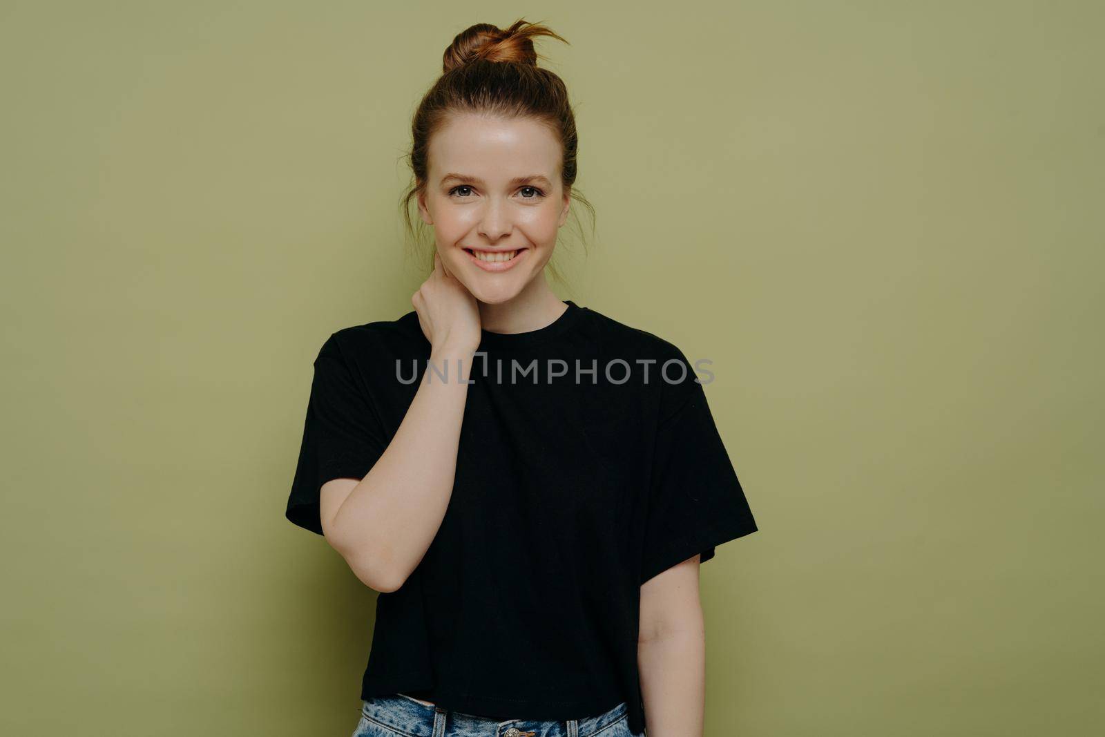 Attractive shy girl in black tshirt with hair in bun doesnt know what to say, being hesitant in decision, holding neck with hand while standing against green wall and expressing positive emotions