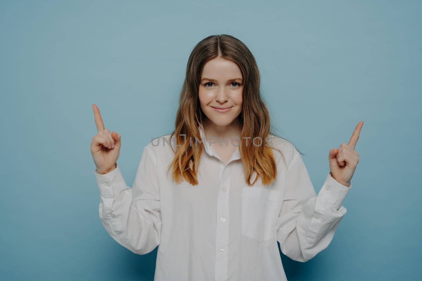 Beautiful teenage girl in white shirt with smug smile and satisfied emotion on face pointing up with her fingers, indicating at copy space, isolated over blue background. Sale and promotion concept