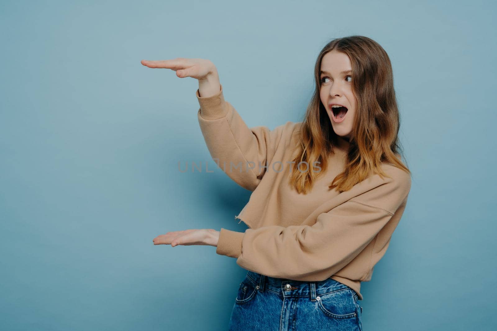 Young shocked brunette woman in beige sweater showing big large size with hands and expressing wow expression while standing light blue background. Promotion and advertising concept