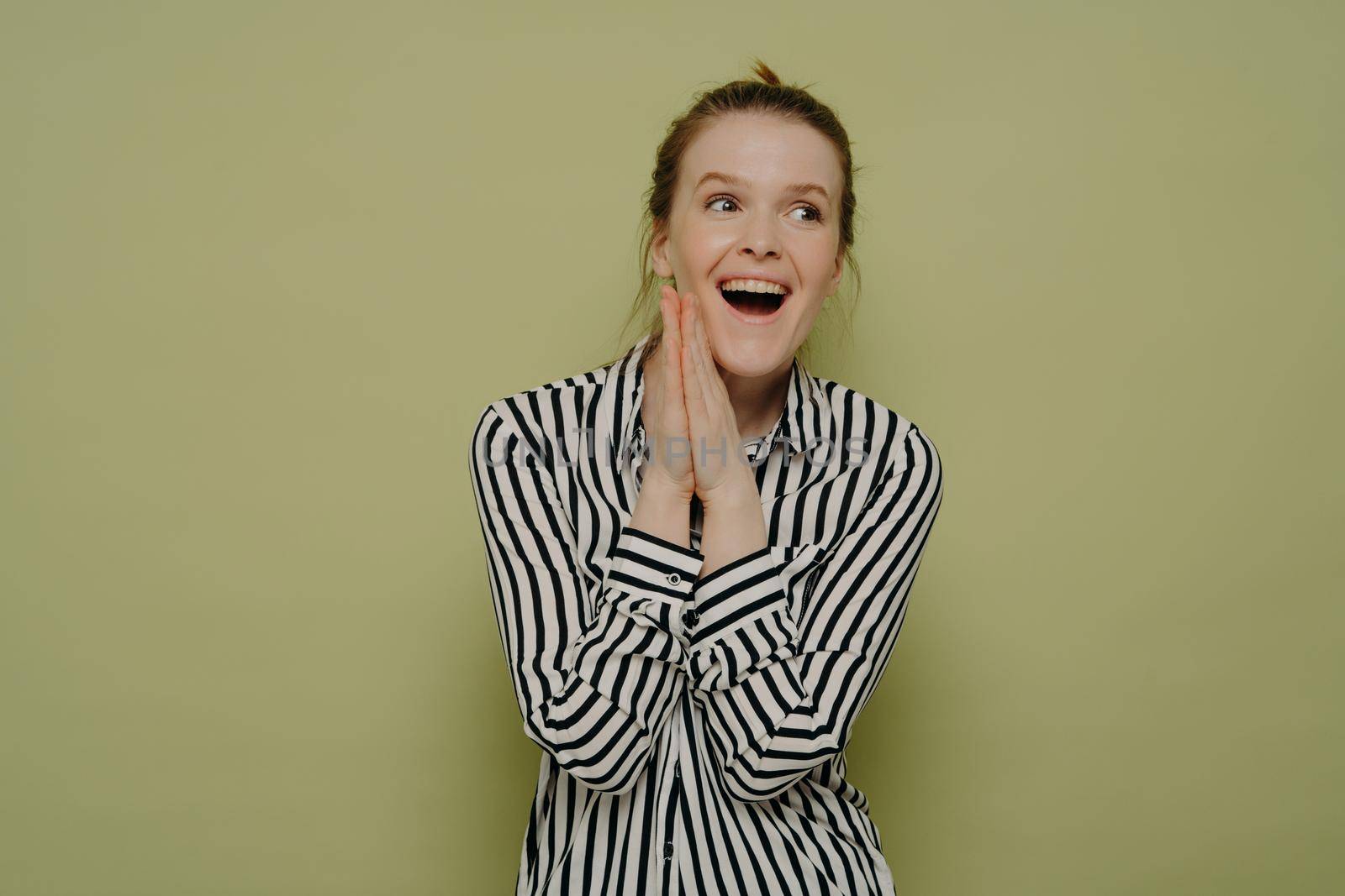 Happy excited lady in white shirt with black stripe with wide open mouth, cant contain her happiness, rejoicing after hearing good news with hands next to face, posing in front of green wall