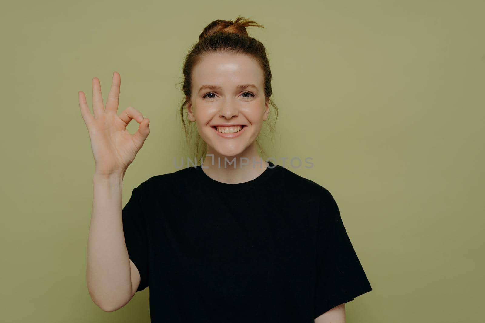 Happy smiling teenage girl with wide smile in black tshirt and hair in bun displaying okay gesture or ok sign with hand, everything going along smoothly, standing in front of green wall