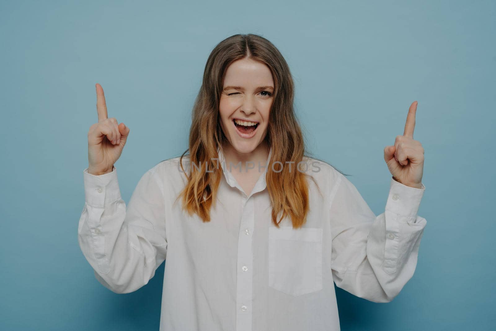 Look here. Happy teenage girl with wide smile in white shirt winking and pointing upwards with her fingers, standing isolated on blue wall. Advertisement concept