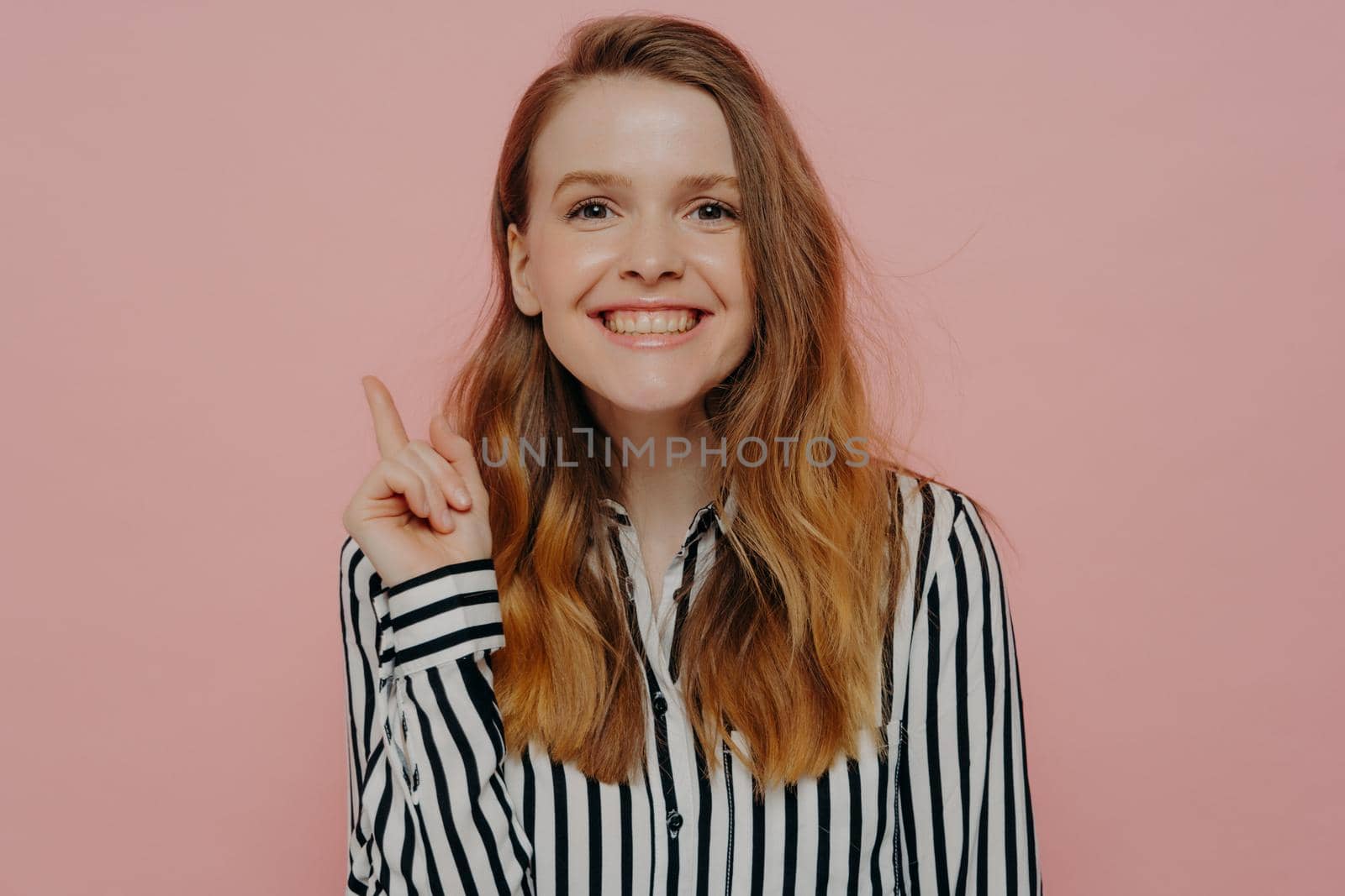 Young smart woman with long hair in white striped shirt, generated new idea, showing it with gesture, teenage girl pointing up with forefinger at copy space and smiling, isolated on pink background