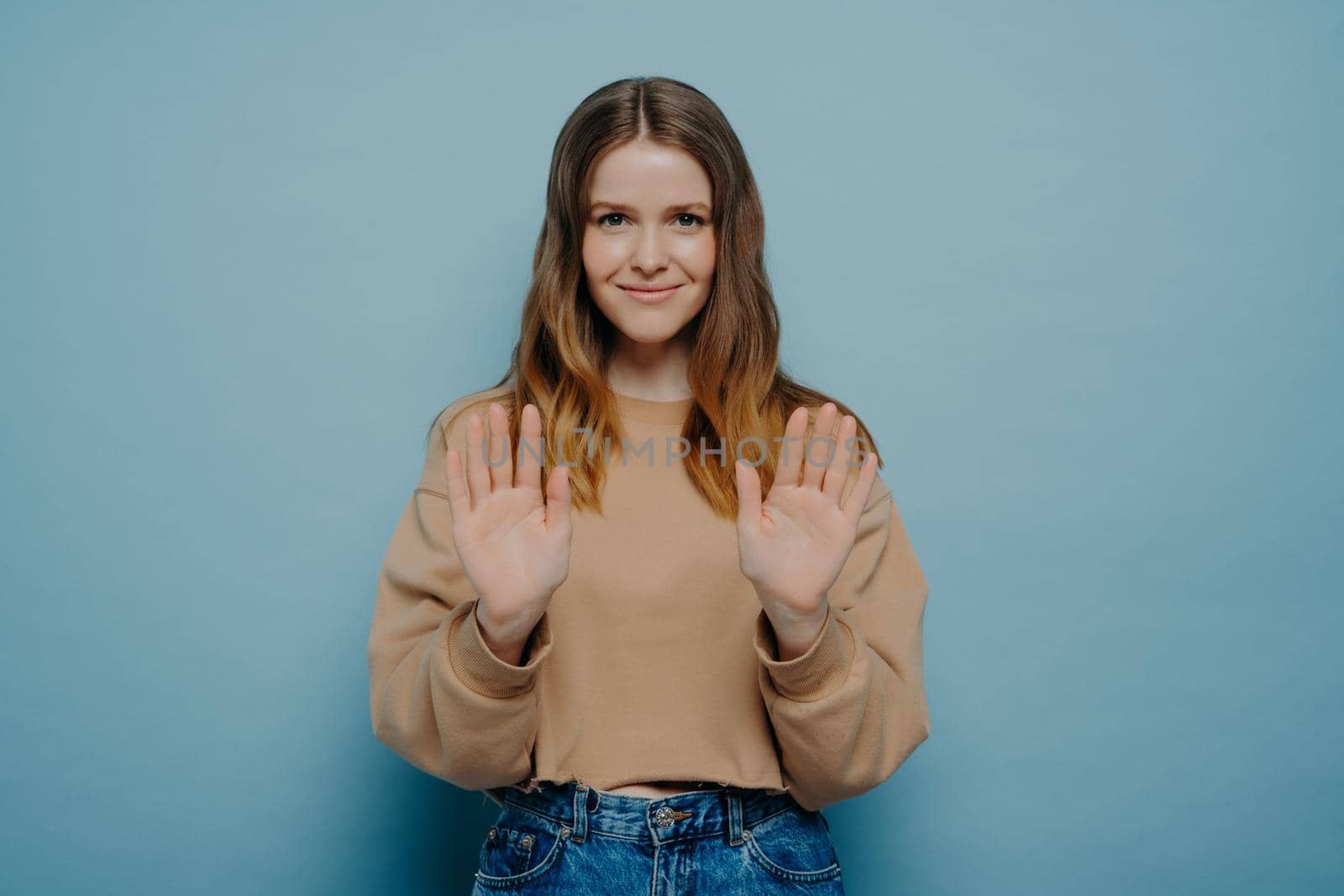Young woman in beige sweater expressing disagreement, showing it by holding hands in front of her, saying no with her gesture, politely refusing to do what she asked on light blue background