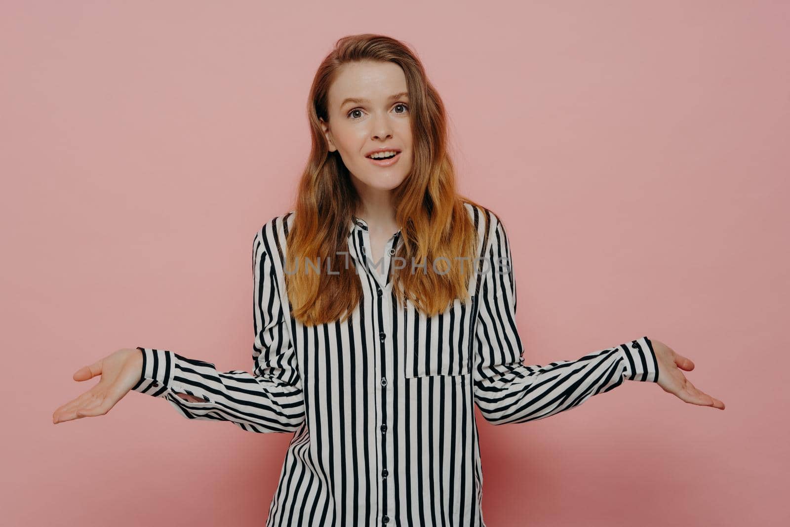 Young woman with long brunette hair wearing white shirt with black stripes cant make important decision or doesnt know how to help, shrugging it off, isolated in front of pink background