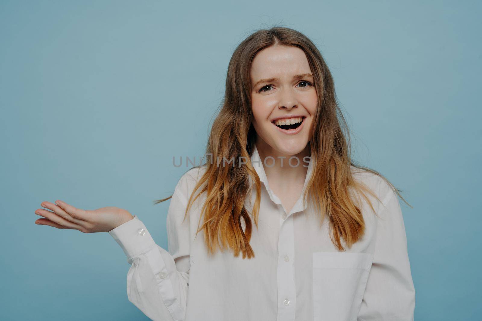 Positive happy female teenager in white shirt presenting something on hand, keeping palm raised while demonstrating or advertising product, standing isolated over blue background