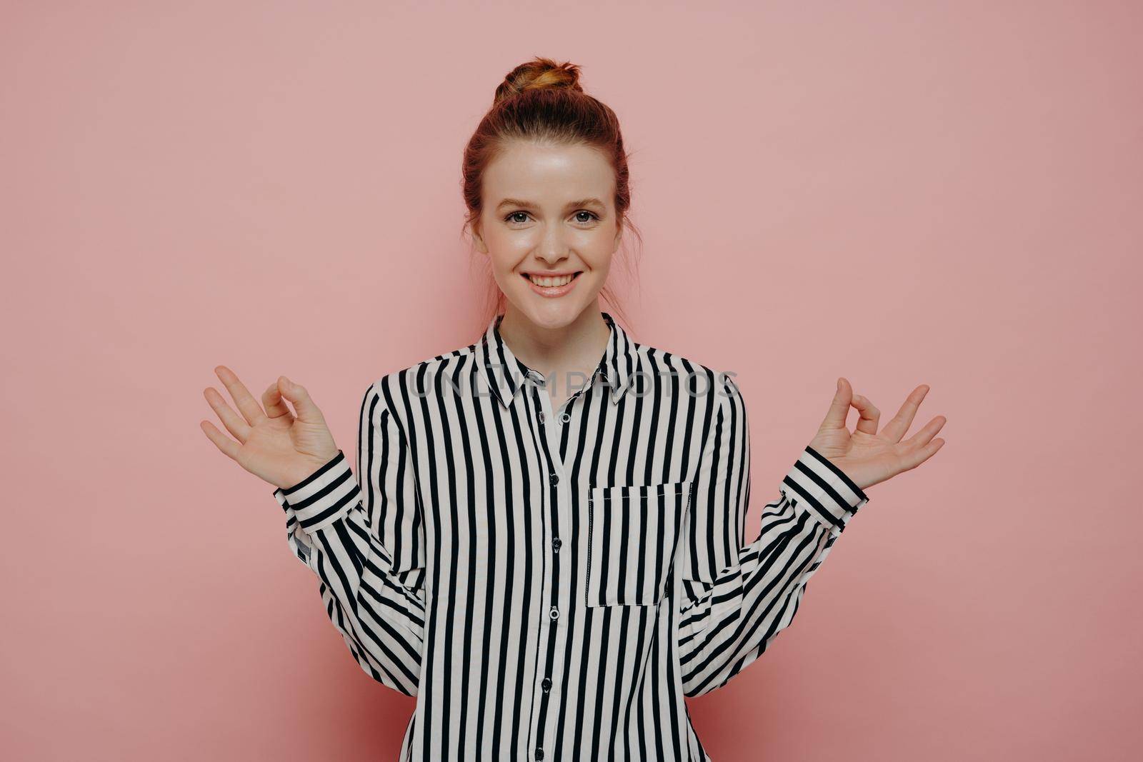 Calm and relaxed young ginger woman with hair in bun in casual wear making okay gesture with both hands or meditating, feeling good while standing alone in front of pink studio background