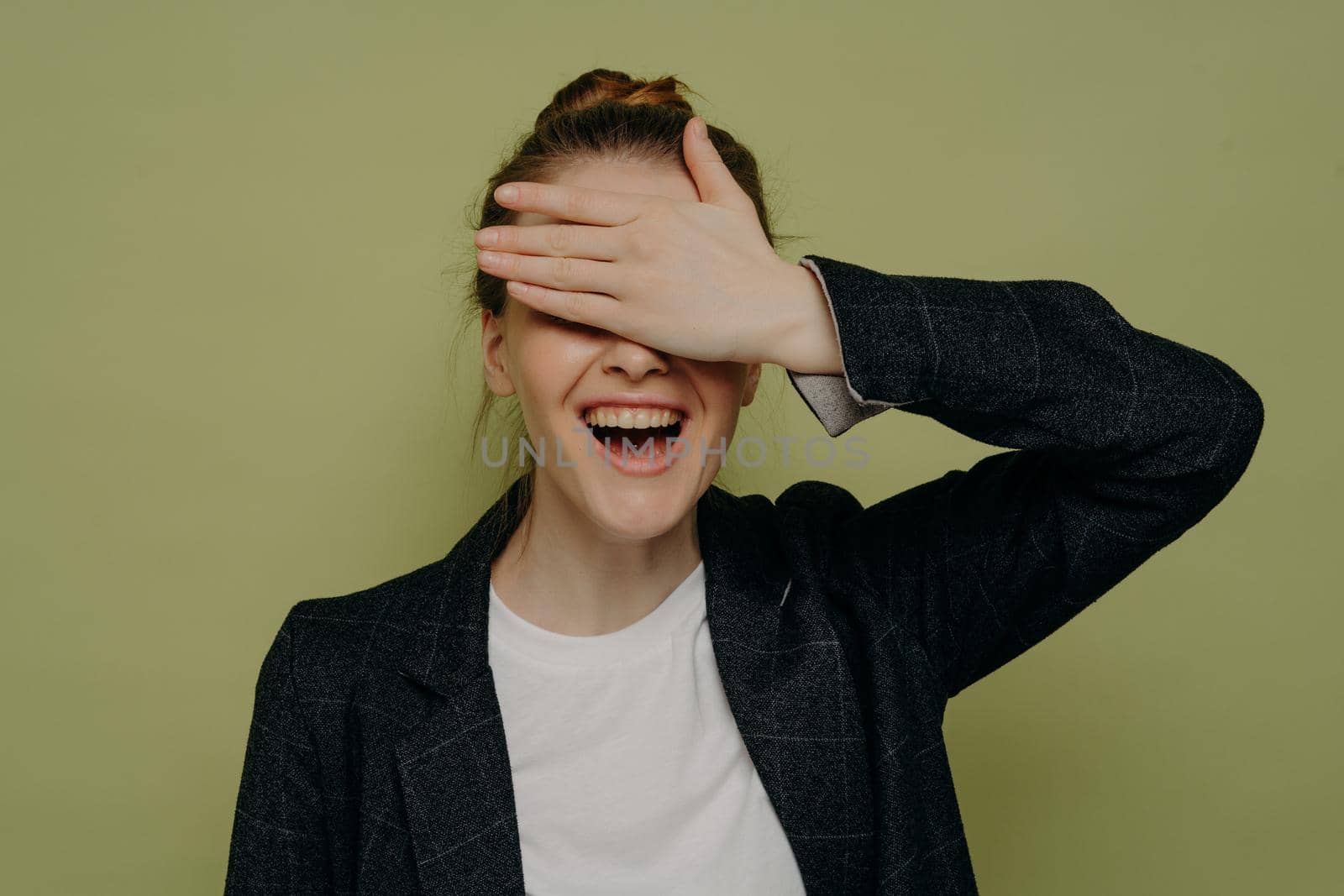 Laughing young female in casual clothes covering eyes with hand while waiting for surprise, smiling teenage girl keeping mouth opened with embarrassed expression, studio shot