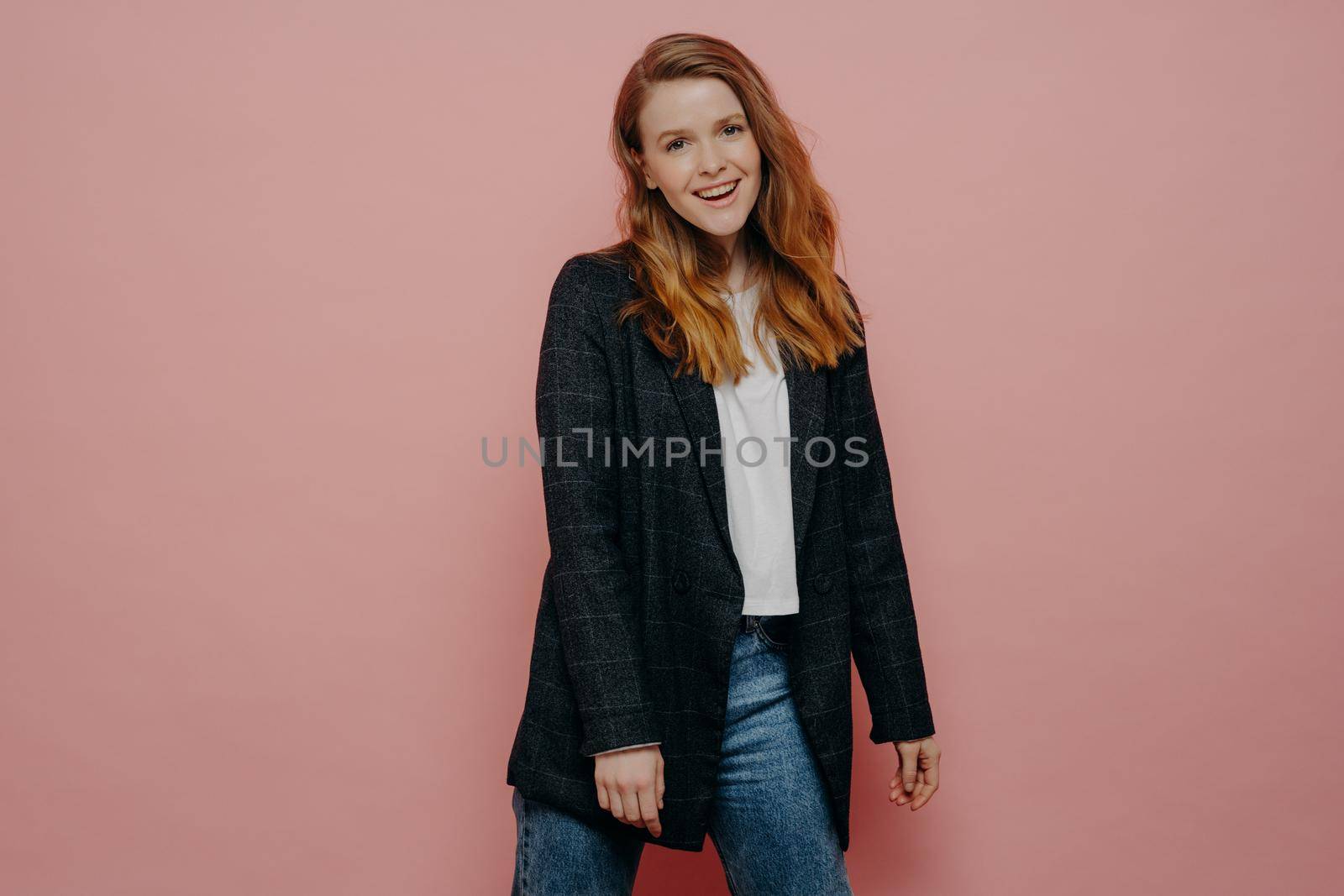 Stylish fashionable young woman wearing plaid blazer and jeans posing in studio, feeling carefree and expressing positive emotions while posing isolated over pink wall background. Happy people concept