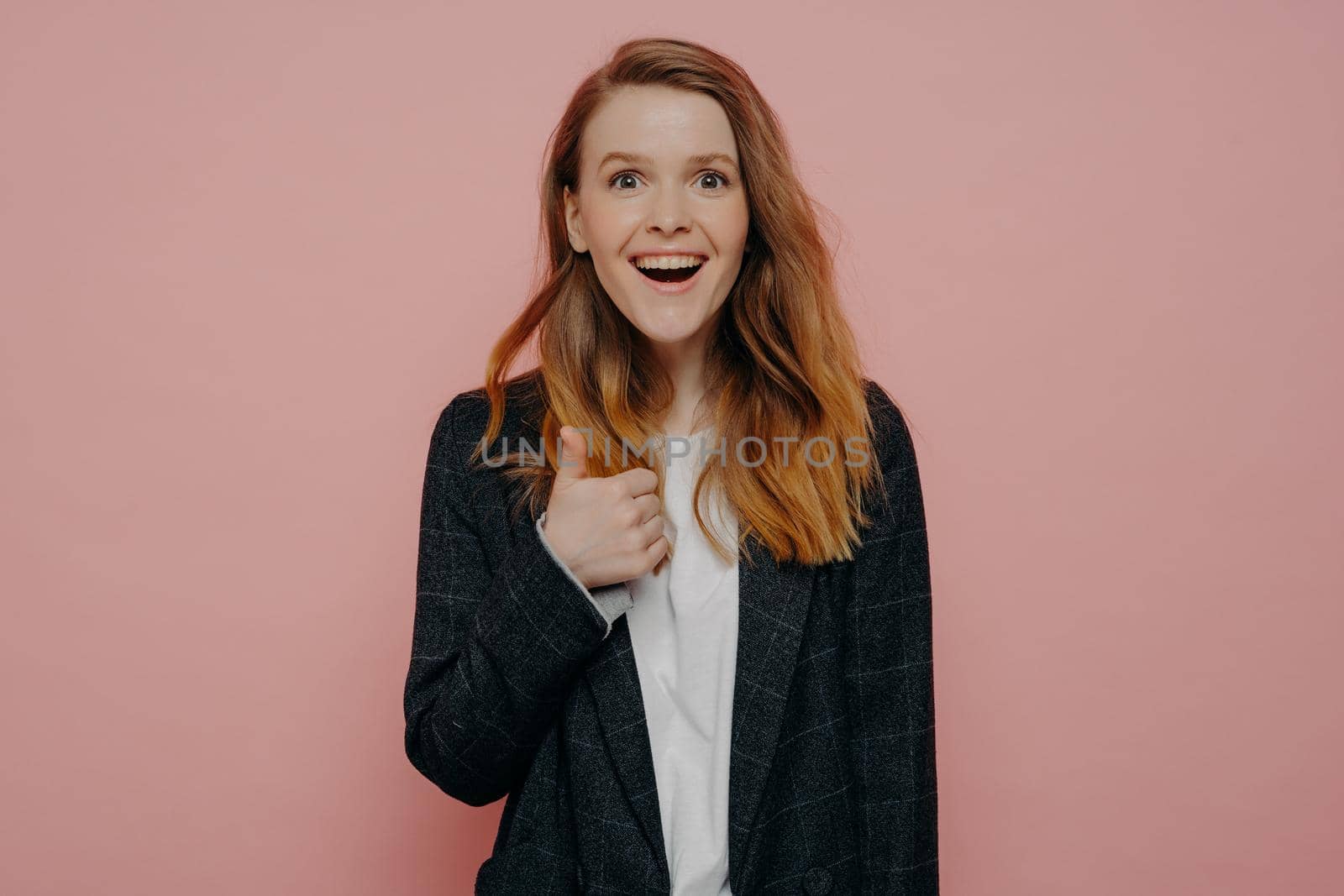 Super. Portrait of excited young redhead woman wearing casual clothes showing thumb up, gesture of approval. looking at camera with movement and keeping moth open. Studio shot