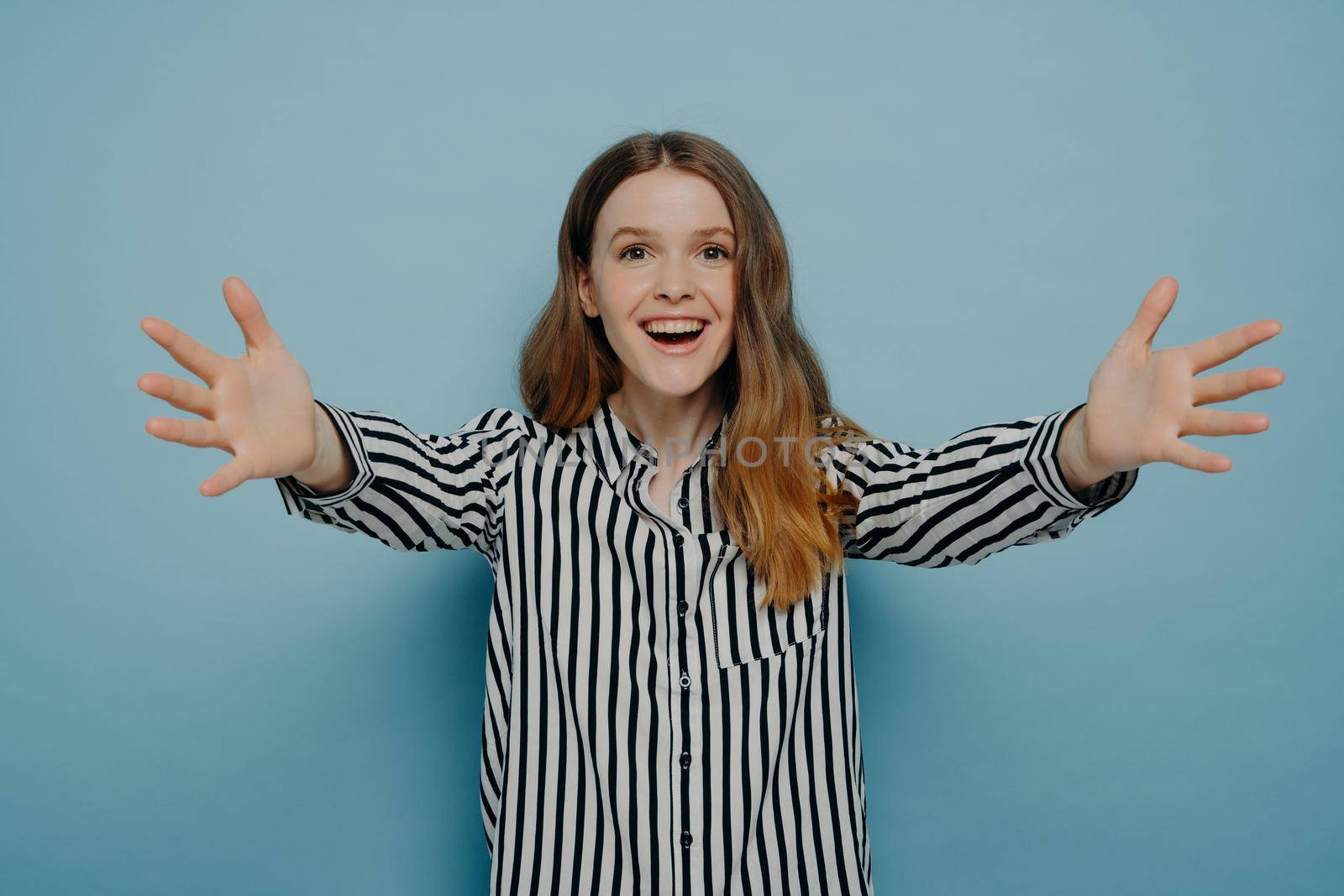 Come here. Young friendly teenage girl giving warm hug to friend while posing against blue wall, looking at camera joyfully and smiling, expressing love and positiveness. Body language concept