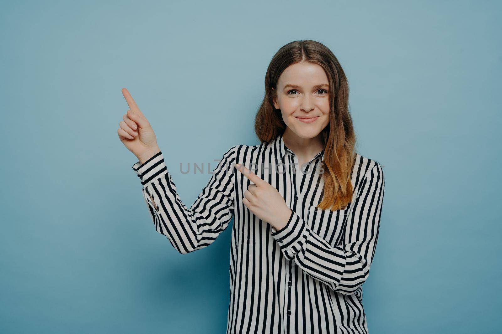 Positive young caucasian female dressed in striped shirt pointing up at at upper left corner with forefingers and smiling at camera, isolated over blue background. Advertisement and promotion