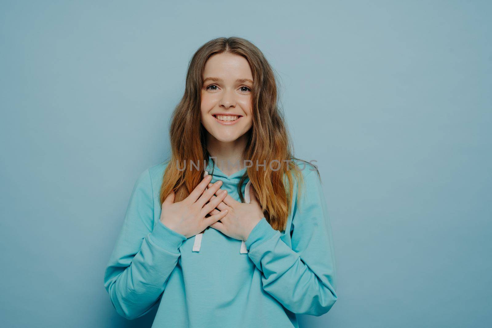 Cute female holding hands on chest and expressing happiness, excited teenage girl dressed in casual wear looking at camera with love and gratitude while standing isolated on blue studio background