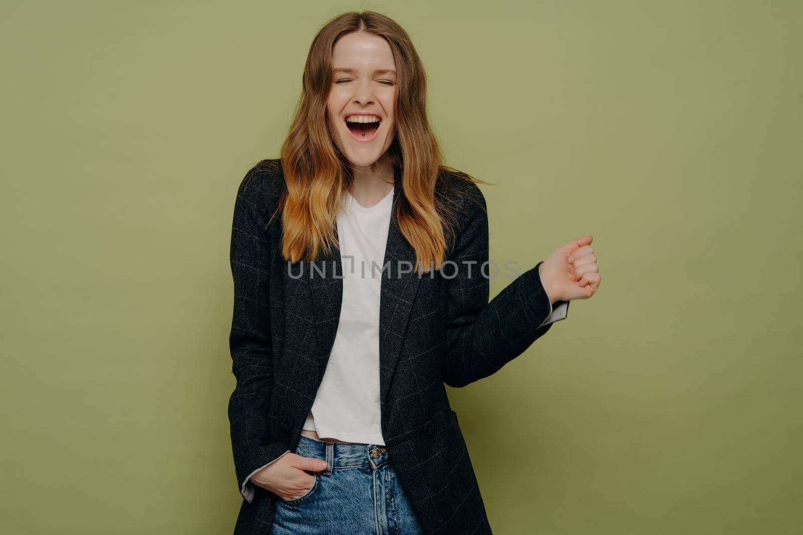 Excitement. Studio shot of surprised and happy young woman with wavy brown hair posing in dark formal jacket, white top and jeans on green background with one hand in pocket. Personal achievements