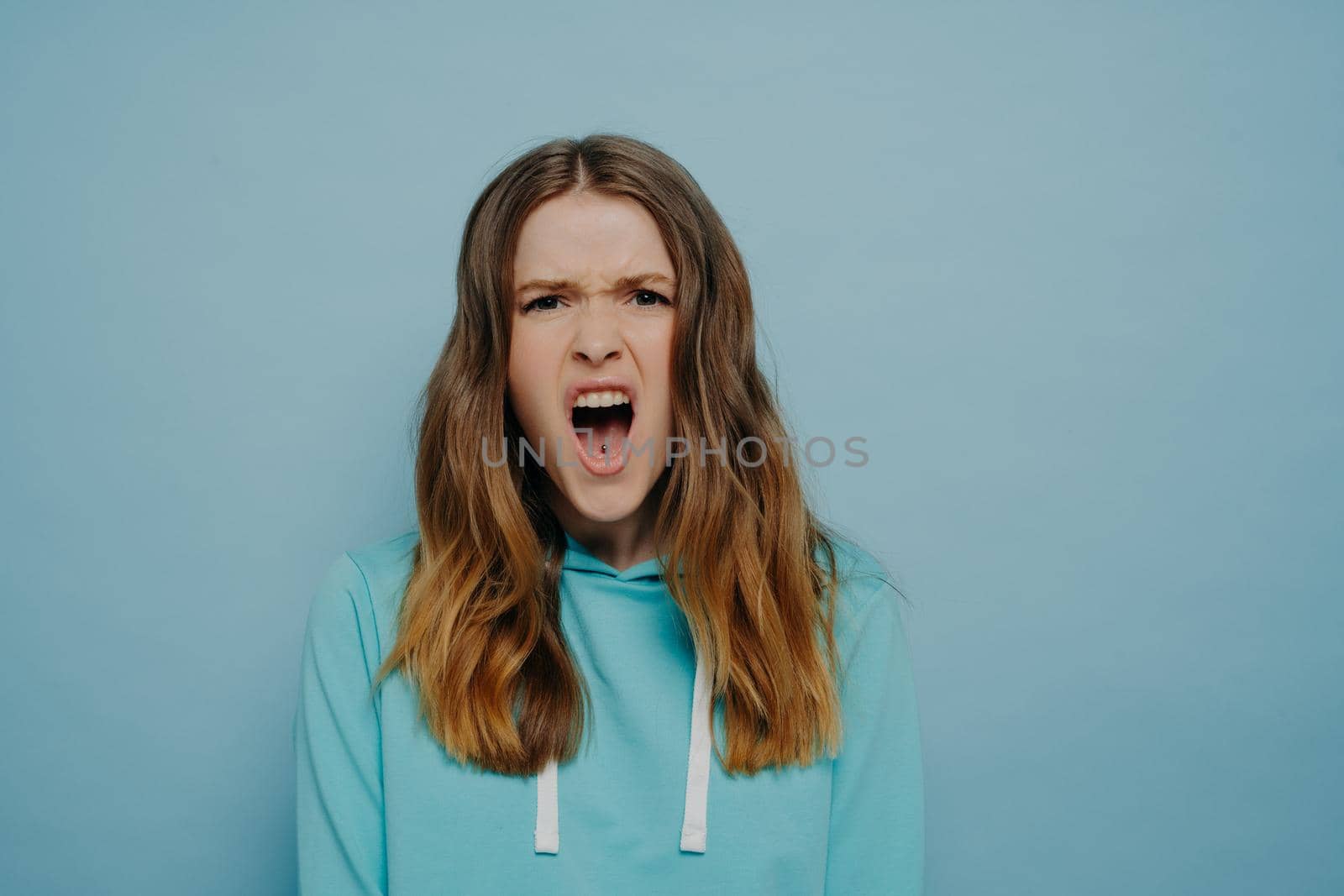 Emotional teenage girl expressing anger and disbelief while looking at camera with open mouth and eyebrows pulled close together, standing against blue background. Negative human emotions concept