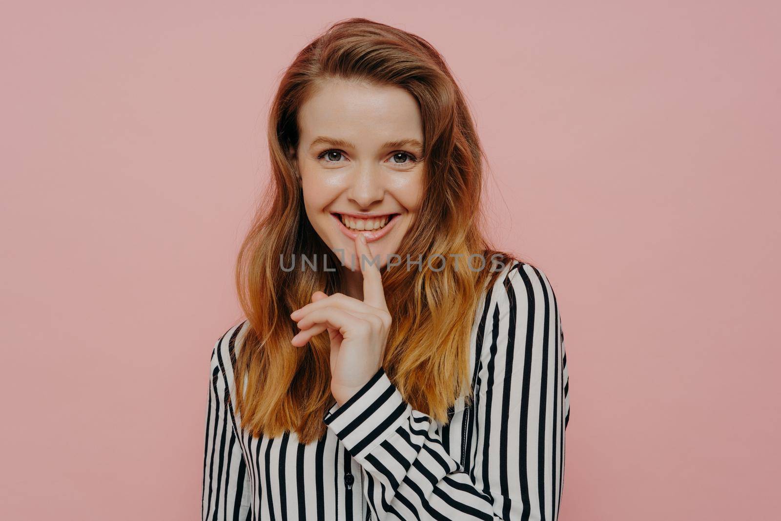 Beautiful happy young female in stripy black and white blouse touching lips with pointer finger while posing in studio with light pink background