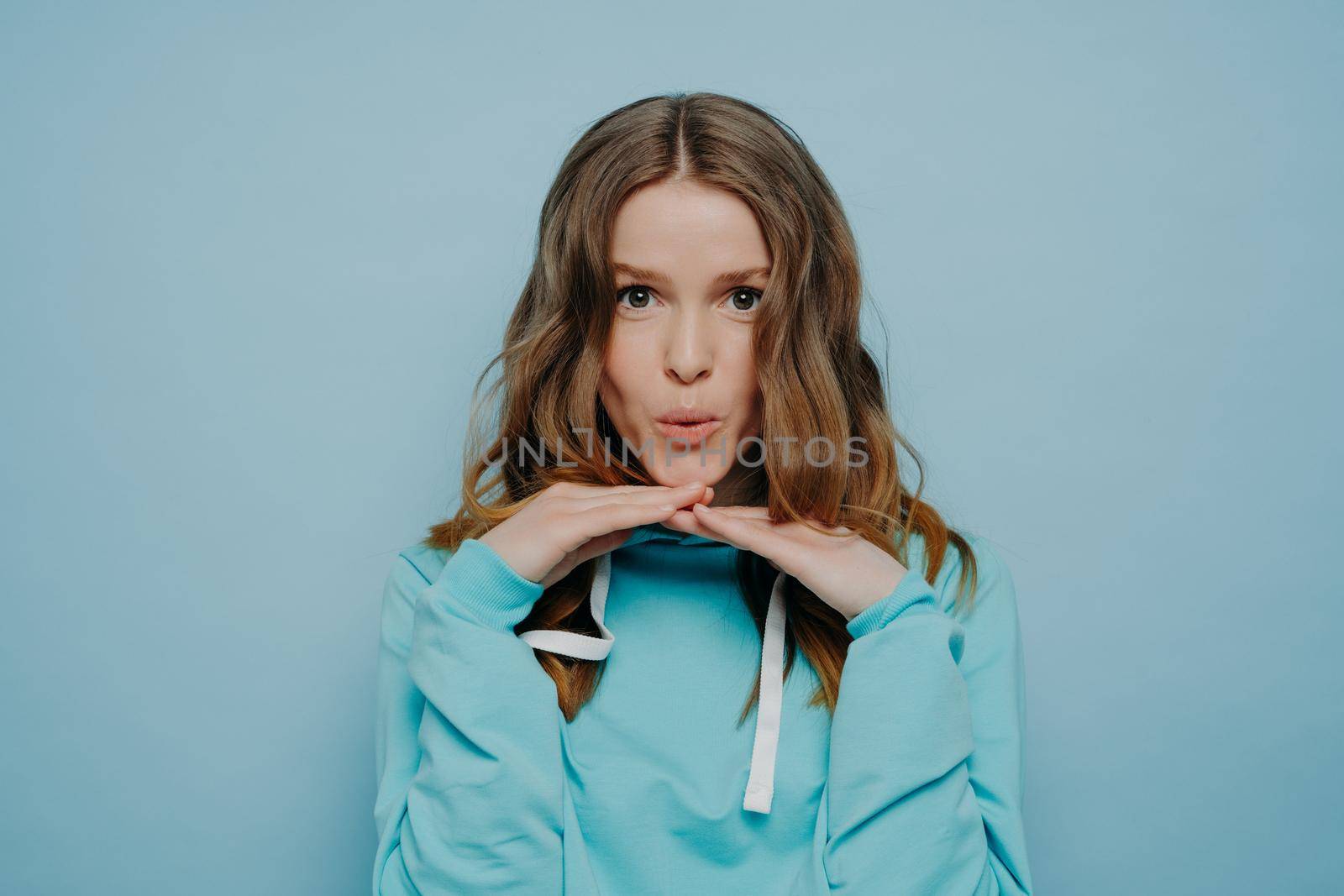 Pretty young teenage girl with wavy brown hair and widely open dark eyes touching her chin with fingertips expressing amusement standing on light blue background. Human emotions concept