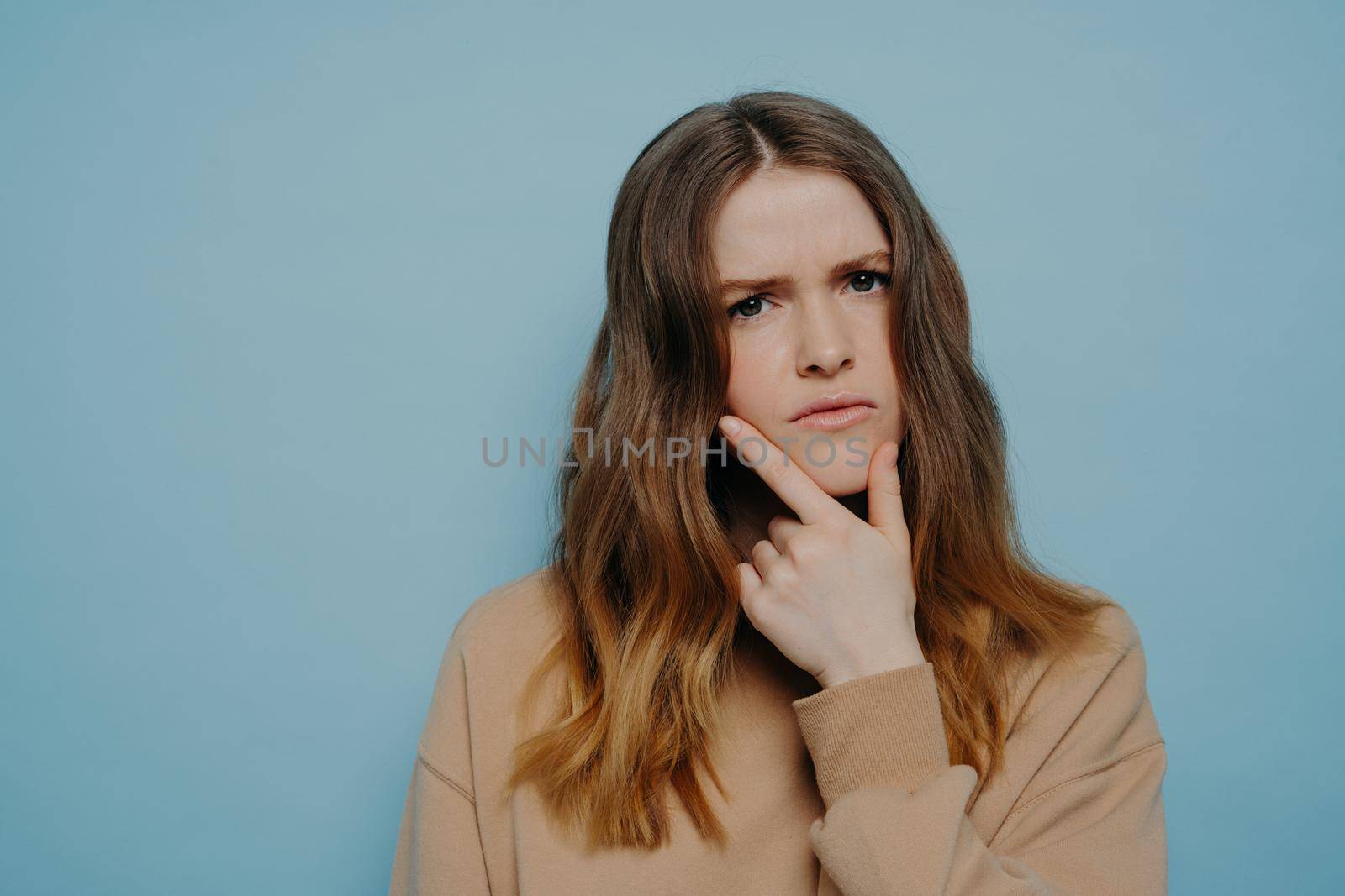 Puzzled young girl with thick wavy ombre hair demonstrating concern and interest while holding hand at chin wearing casual light brown sweater and standing against blue studio background
