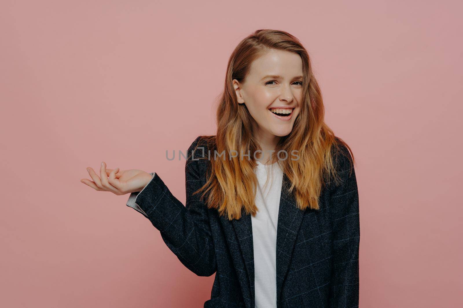 Excited young woman with tipping hand shrugging shoulders standing against light pink background in dark formal jacket, cute girl with wavy ginger medium length hair laughing