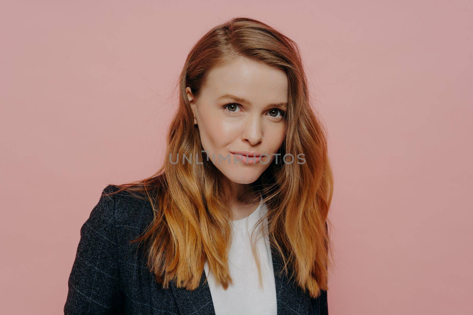 Portrait of beautiful young woman with ginger hair looking at camera confidently, wearing dark formal jacket and white top posing isolated against pink studio wall. Human emotions concept