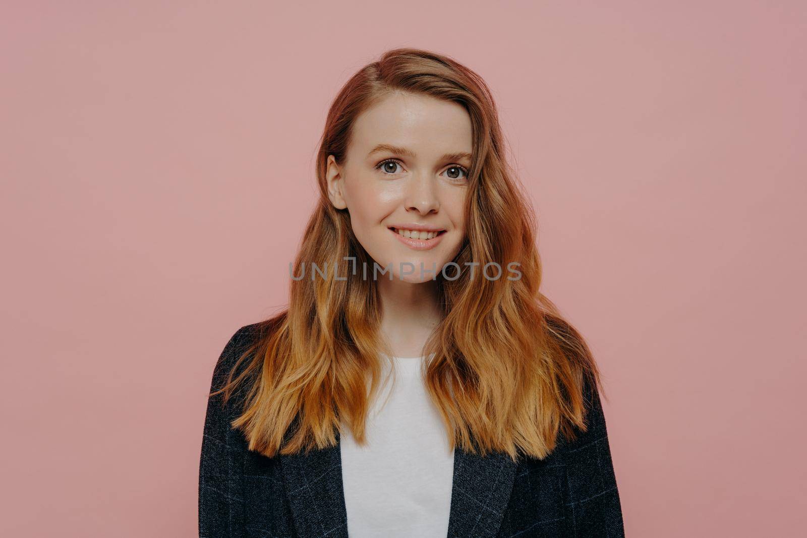 Photo of pretty smiling female with ginger hair without makeup looking at camera wearing dark formal jacket and white top, happy teenage girl posing isolated over pink studio background