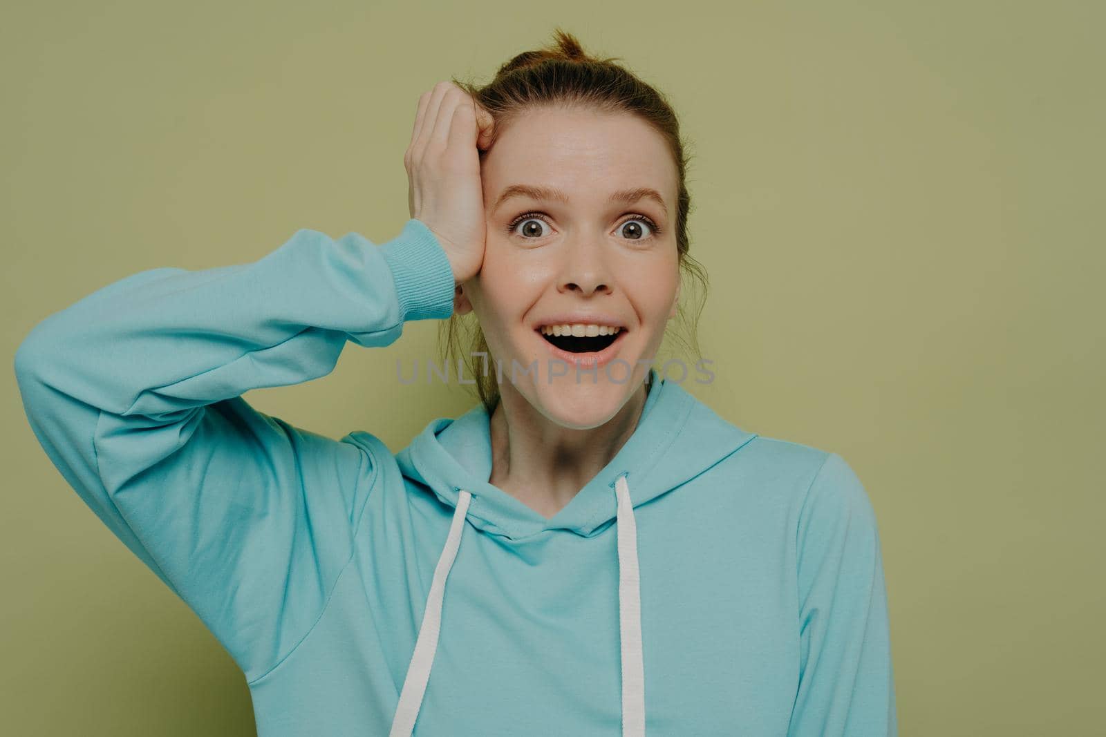 Surprised young woman without makeup demonstrating shock and excitement holding hand at head looking at camera wearing casual blue hooded sweater while posing in studio. Human emotions concept