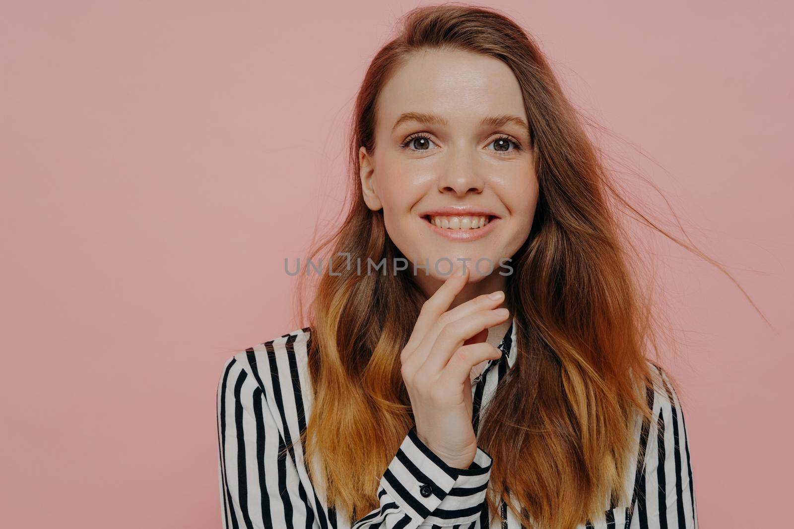 Photo of cute smiling young female with thick wavy ginger hair in formal striped black and white blouse standing over light pink studio background while touching her chin. Happiness concept