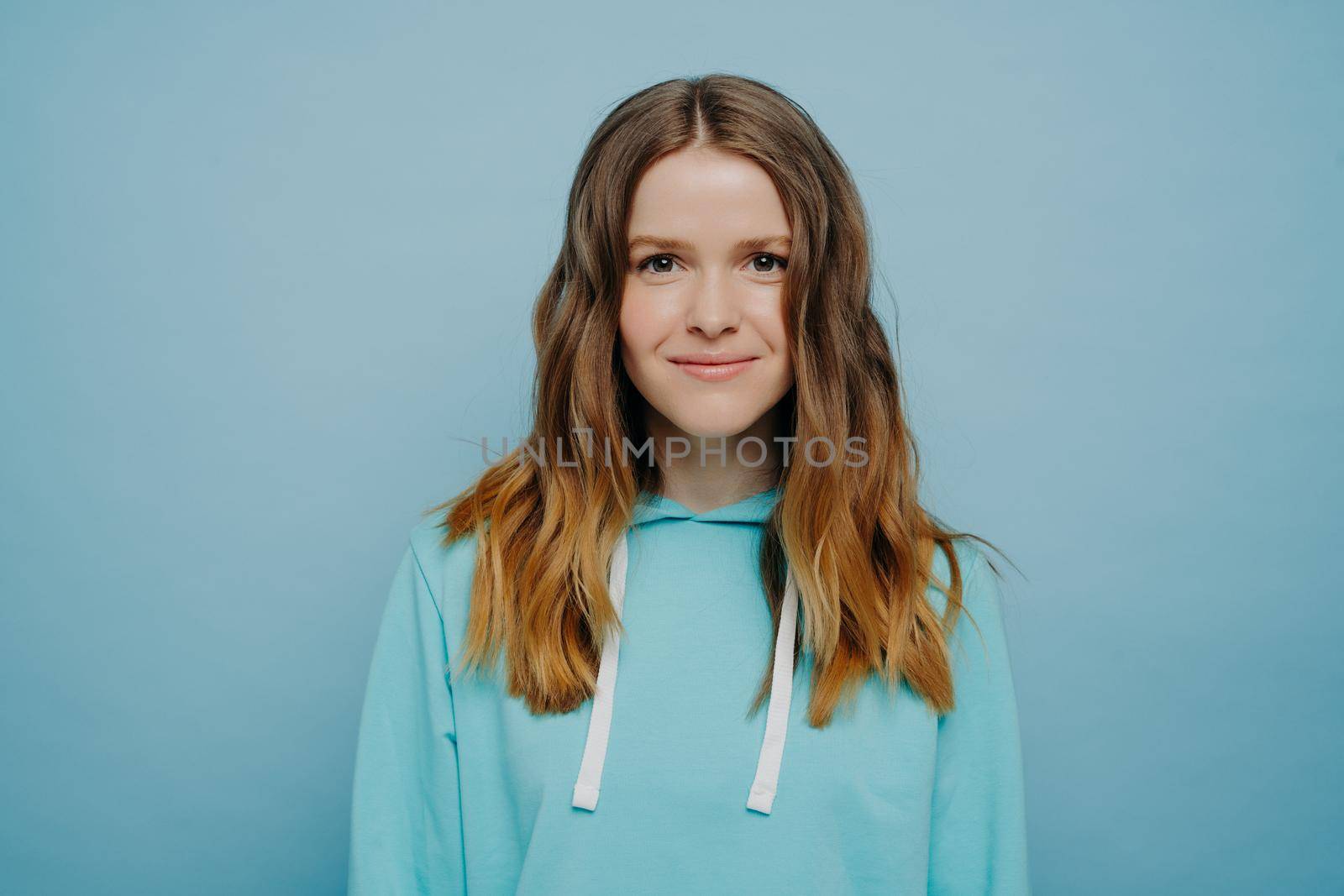 Young attractive girl with wavy ombre hairstyle smiling while looking at camera in comfortable casual hooded sweatshirt standing against light blue background. Medium shot