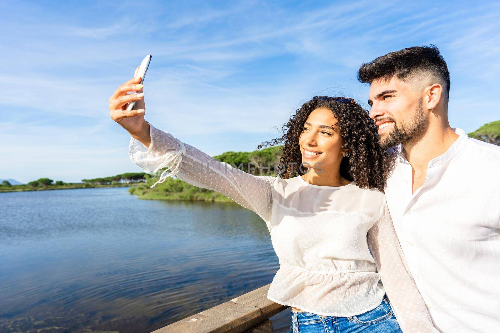 Happy couple in love making a selfie on vacation on a sea resort at sunset. Young Afro-American woman using a smartphone for a self-portrait with her boyfriend on a lake. Millennials living nature by robbyfontanesi