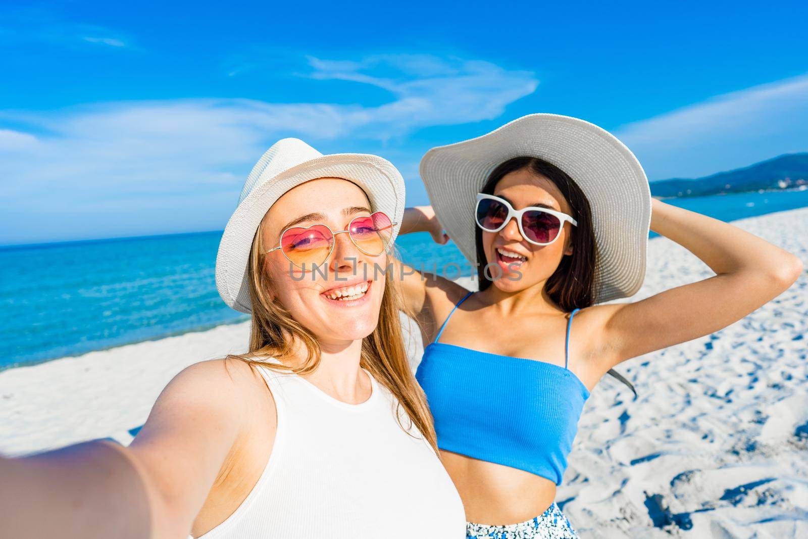 Two girls taking a self-portrait with cell wearing blue casual summer clothes and funny sunglasses on vacation at tropical beach resort. Beauty young women couple enjoying travel using smartphone