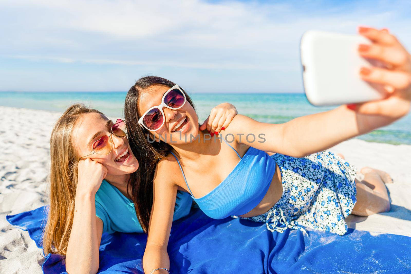 Two cute girls lying on white sand taking a self-portrait with cell wearing blue casual summer clothes and funny sunglasses on vacation at tropical beach resort with blue ocean sea on background