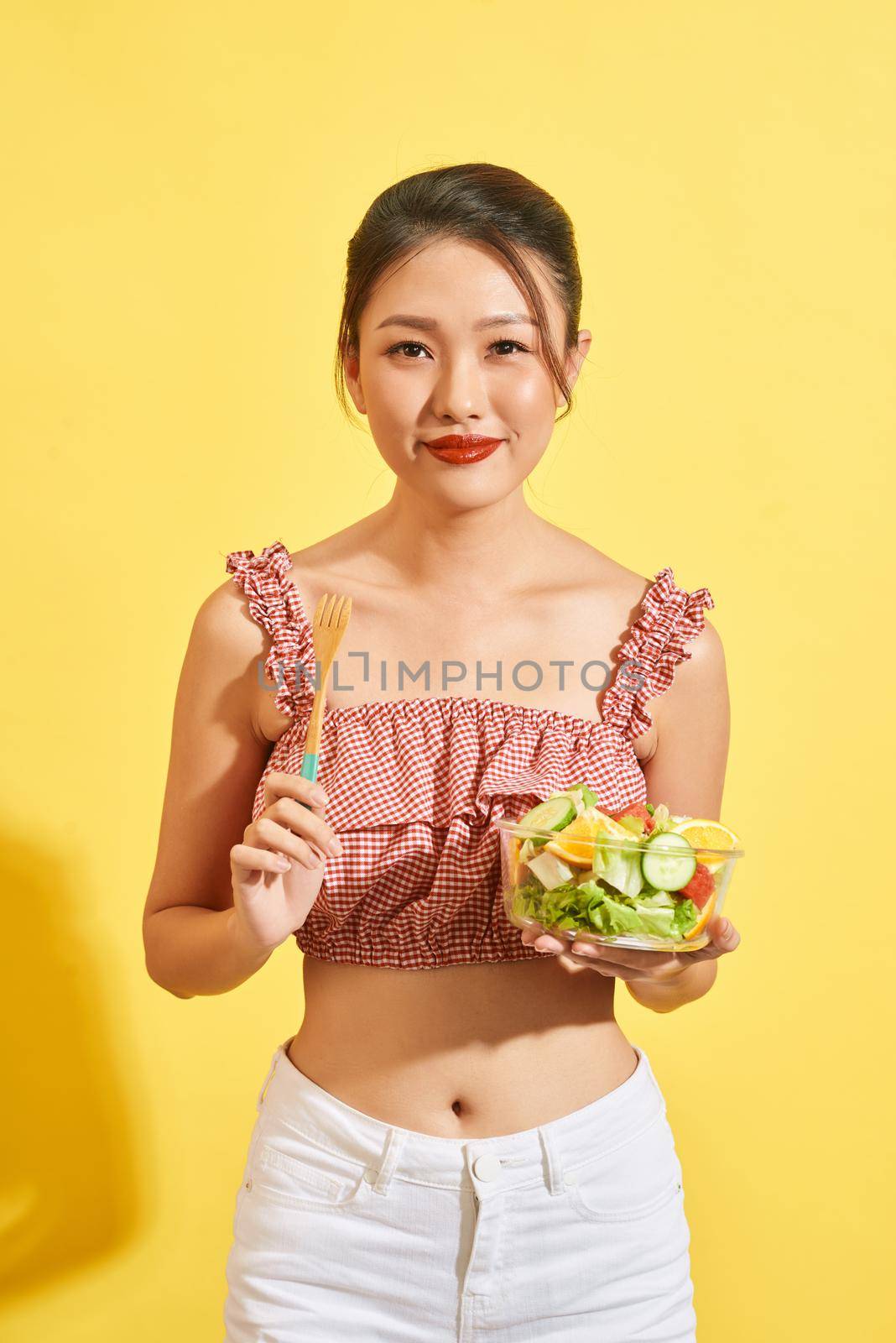 young Asian woman smiling and holding vegetable and salad on yellow background