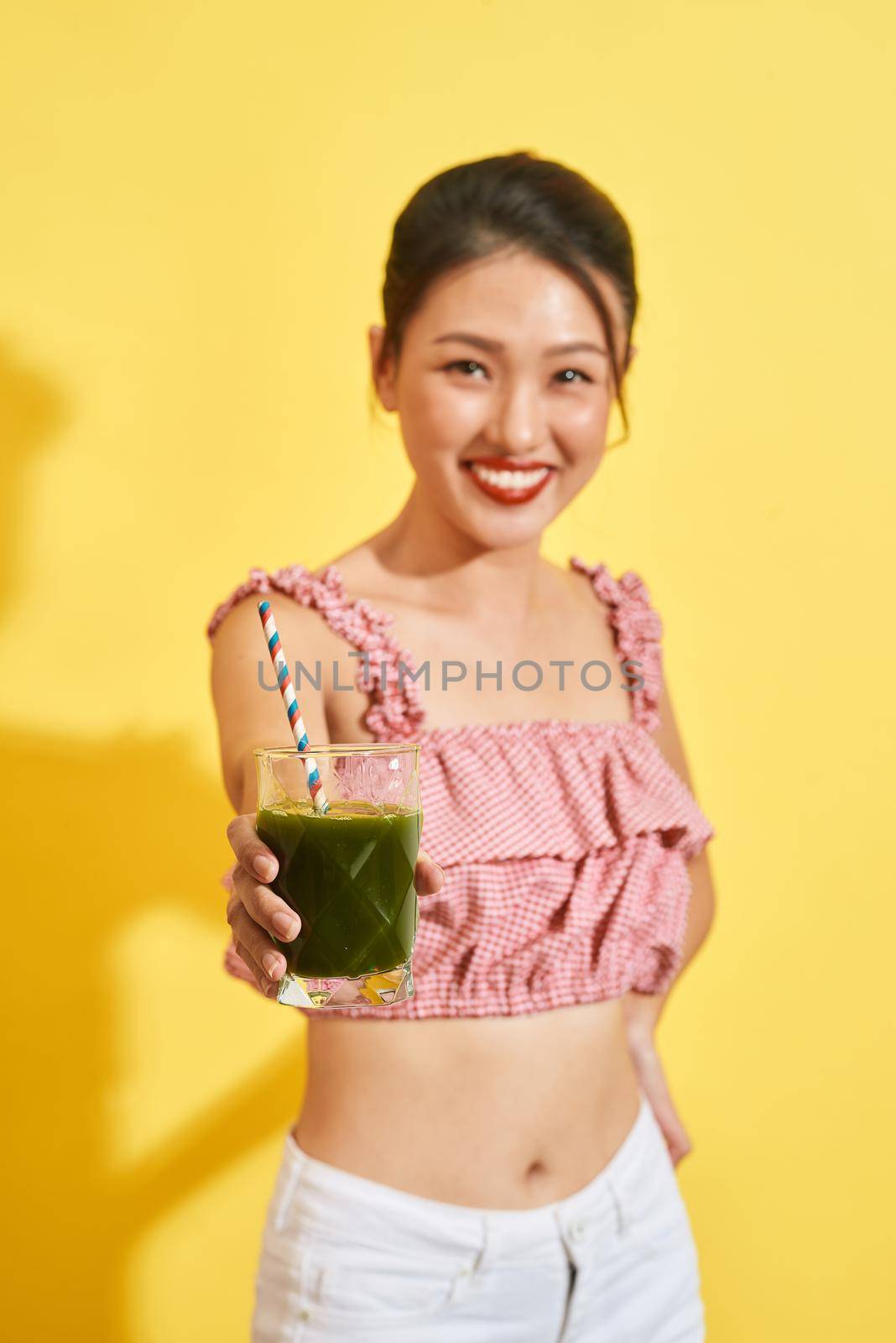 Smiling young asian woman holding green fresh vegetable juice or smoothie from glass 