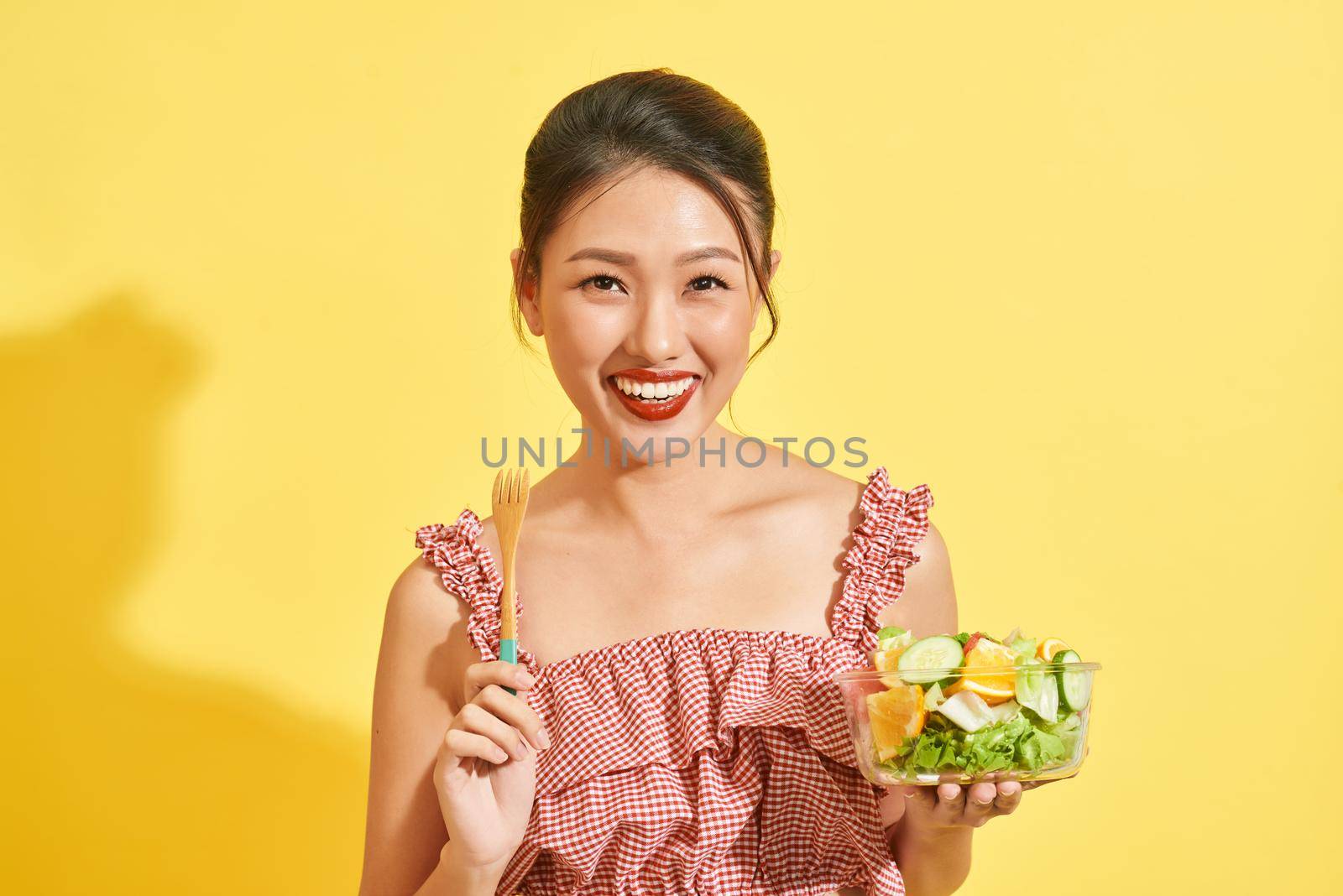 young Asian woman smiling and holding vegetable and salad on yellow background