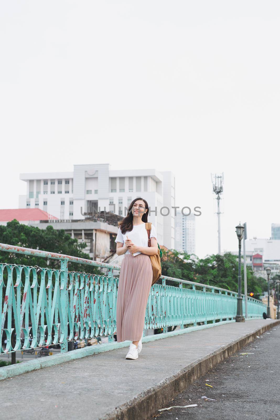 Full length of asian girl walking on a bridge with city view with a book in her hand by makidotvn