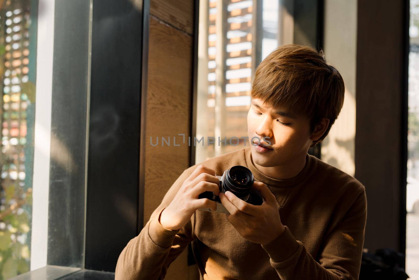 Asian Male professional photographer dressed in casual outfit making setting on display choosing filters on vintage camera and watching photos while resting in coffee shop interior near window