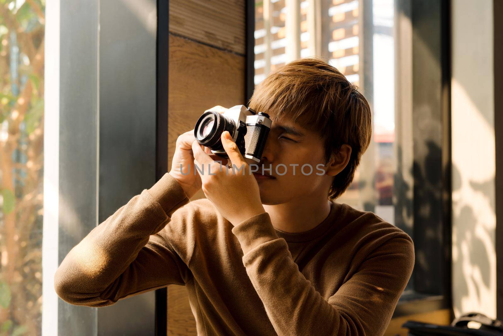 Male amateur photographer in eyeglasses focusing to make photos out of window sitting on windowsill.Young bearded man resting in coworking space and taking pictures for portfolio on vintage camera by makidotvn
