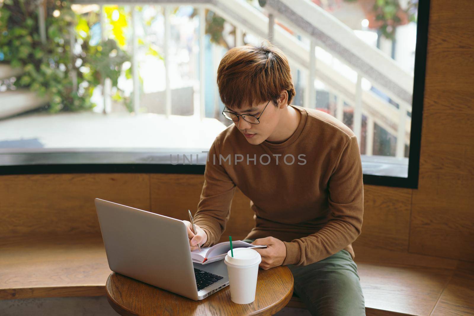 Cheerful male journalist in trendy glasses happy to finishing work on book review rereading written in notebook text before calling to editor to present publication drinking coffee for lunch in cafe by makidotvn