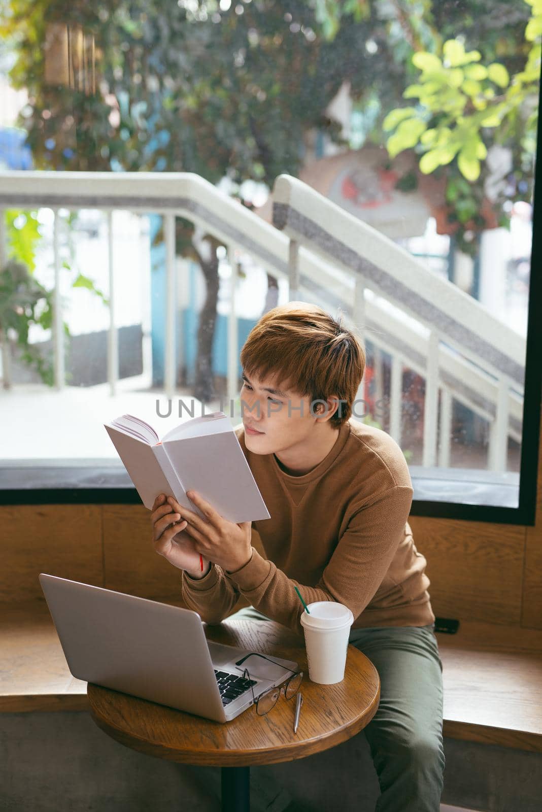 Pensive young asian businessman reading book at desktop with laptop, coffee cup in coffee shop. Knowledge concept