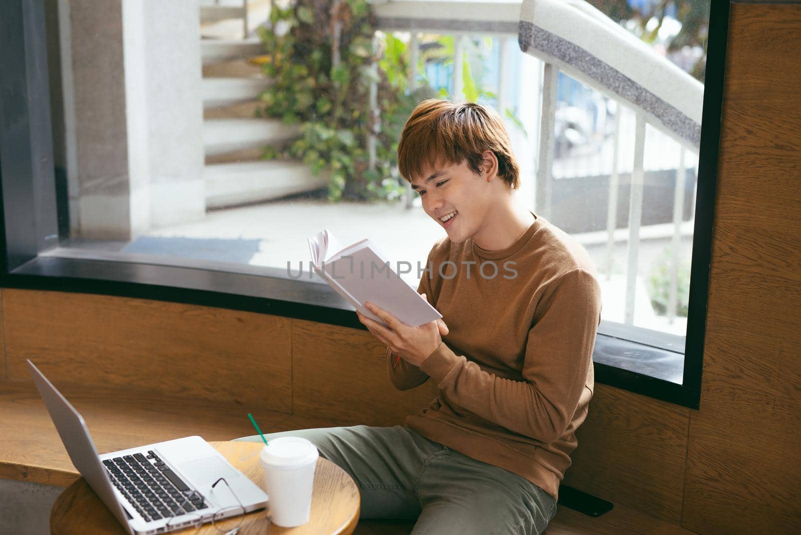 Pensive young asian businessman reading book at desktop with laptop, coffee cup in coffee shop. Knowledge concept by makidotvn