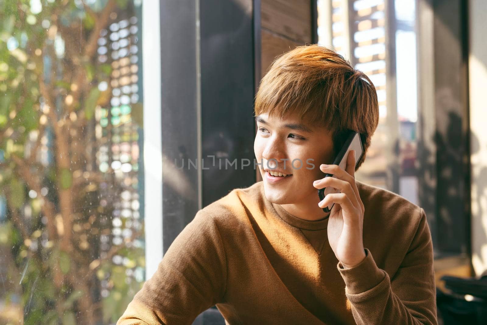 Closeup portrait of smiling young Asian man talking on mobile phone and sitting at empty table with blurred cafe interior in background