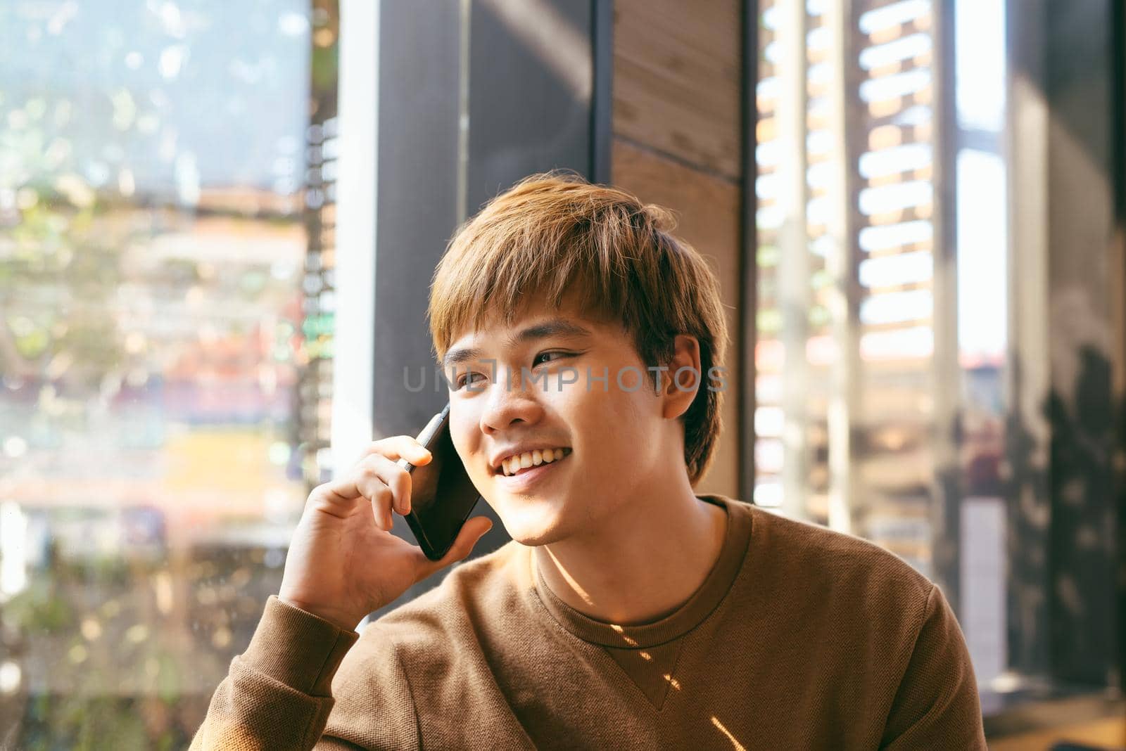 Closeup portrait of smiling young Asian man talking on mobile phone and sitting at empty table with blurred cafe interior in background