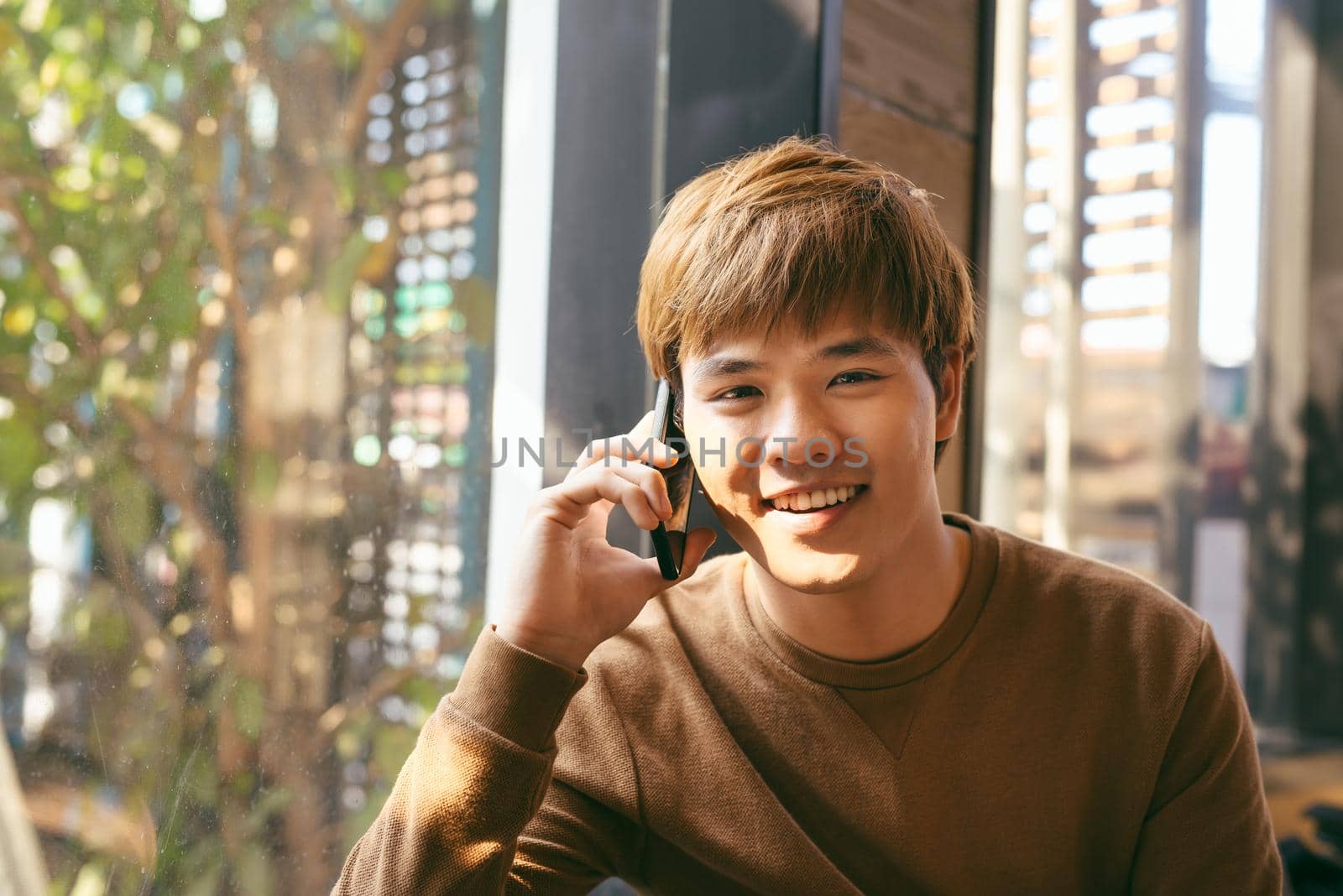 Closeup portrait of smiling young Asian man talking on mobile phone and sitting at empty table with blurred cafe interior in background by makidotvn