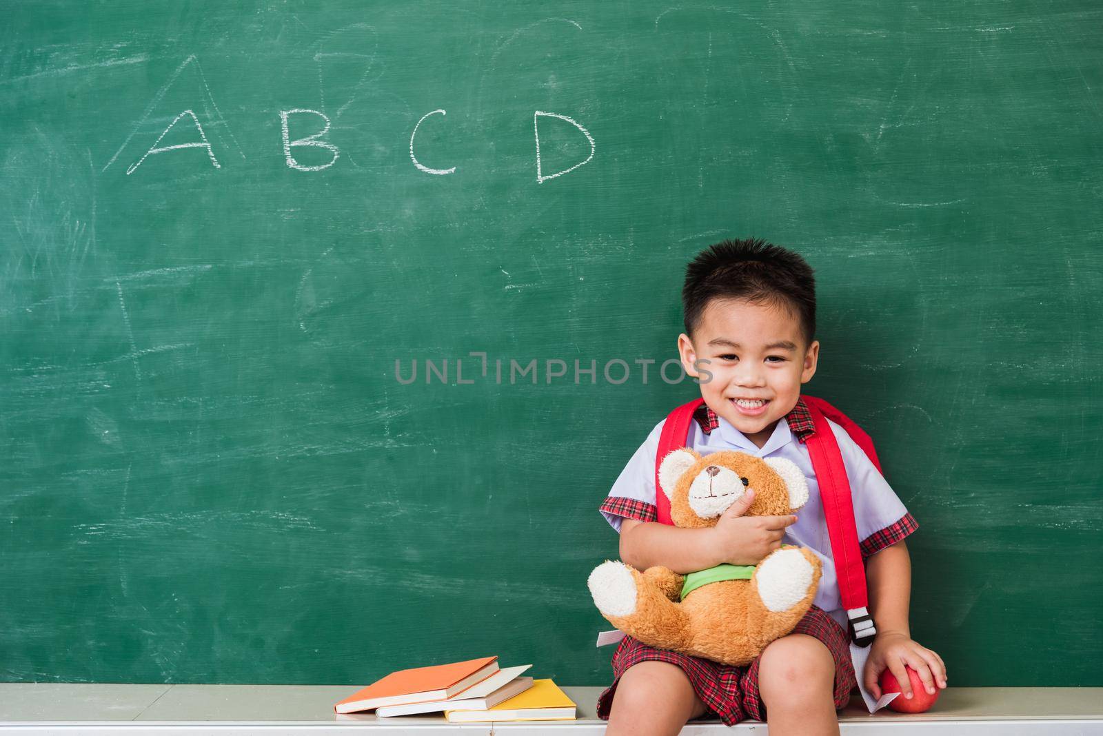 Back to School. Happy Asian funny cute little child boy from kindergarten in student uniform with school bag smiling and hugging teddy bear on green school blackboard, First time to school education