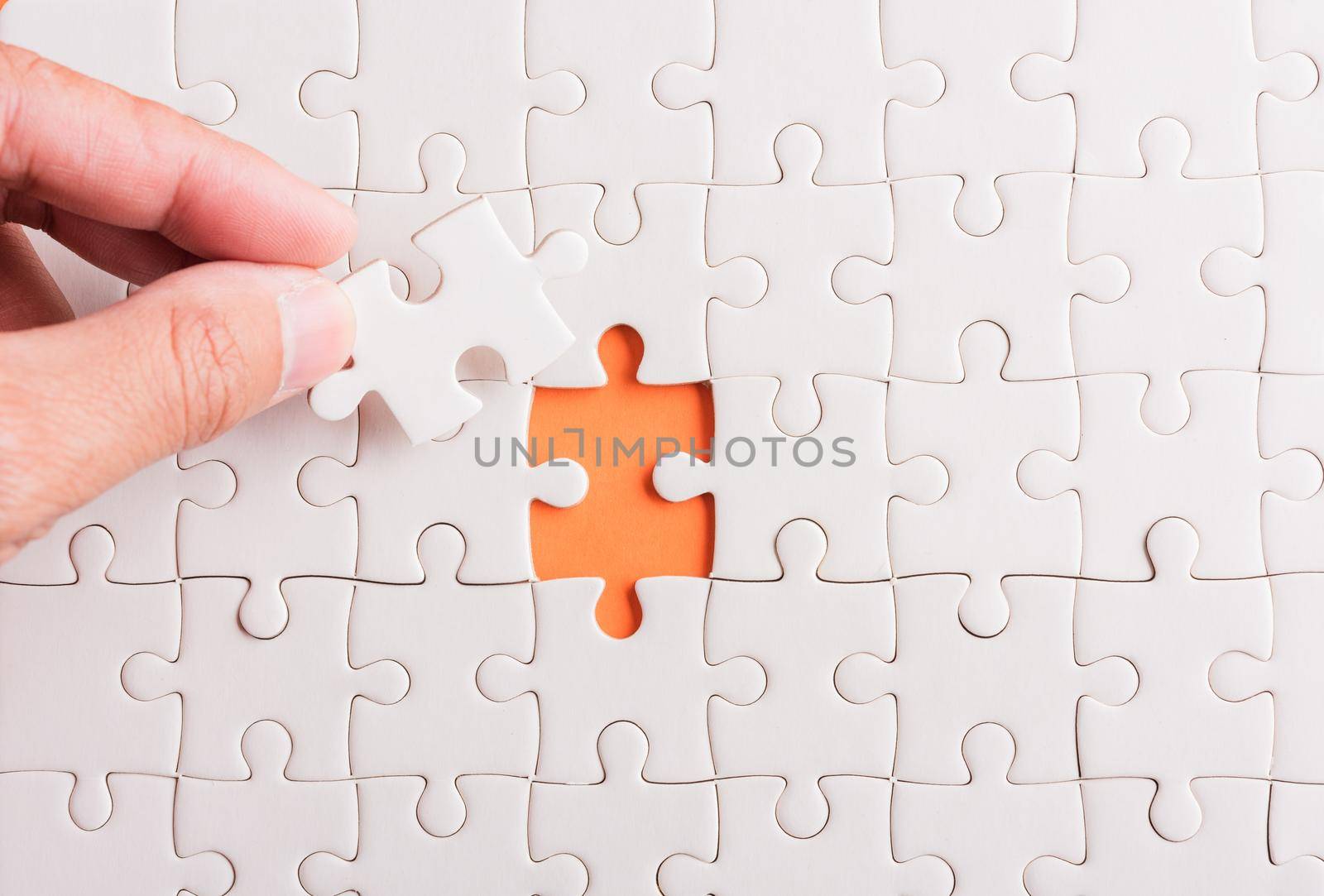 Top view flat lay of hand-holding last piece white paper jigsaw puzzle game last pieces put to place for solve problem complete mission, studio shot on an orange background, quiz calculation concept