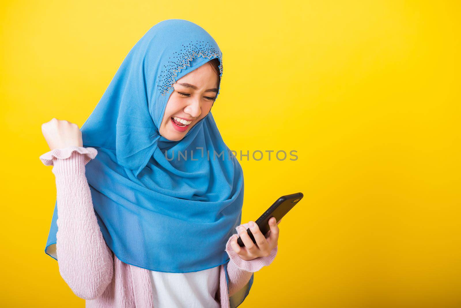 Asian Muslim Arab, Portrait of happy beautiful young woman Islam religious wear veil hijab funny smile she raise hand glad celebrating winning with smart mobile phone isolated on yellow background