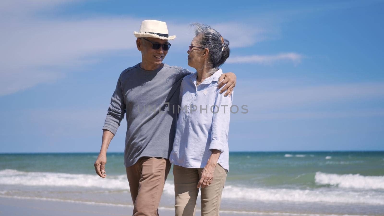 Happy Asian senior man and woman couple holding hands walking from the beach sunny with bright blue sky, Romantic elderly enjoy Travel summer vacation, plan life insurance at retirement couple concept